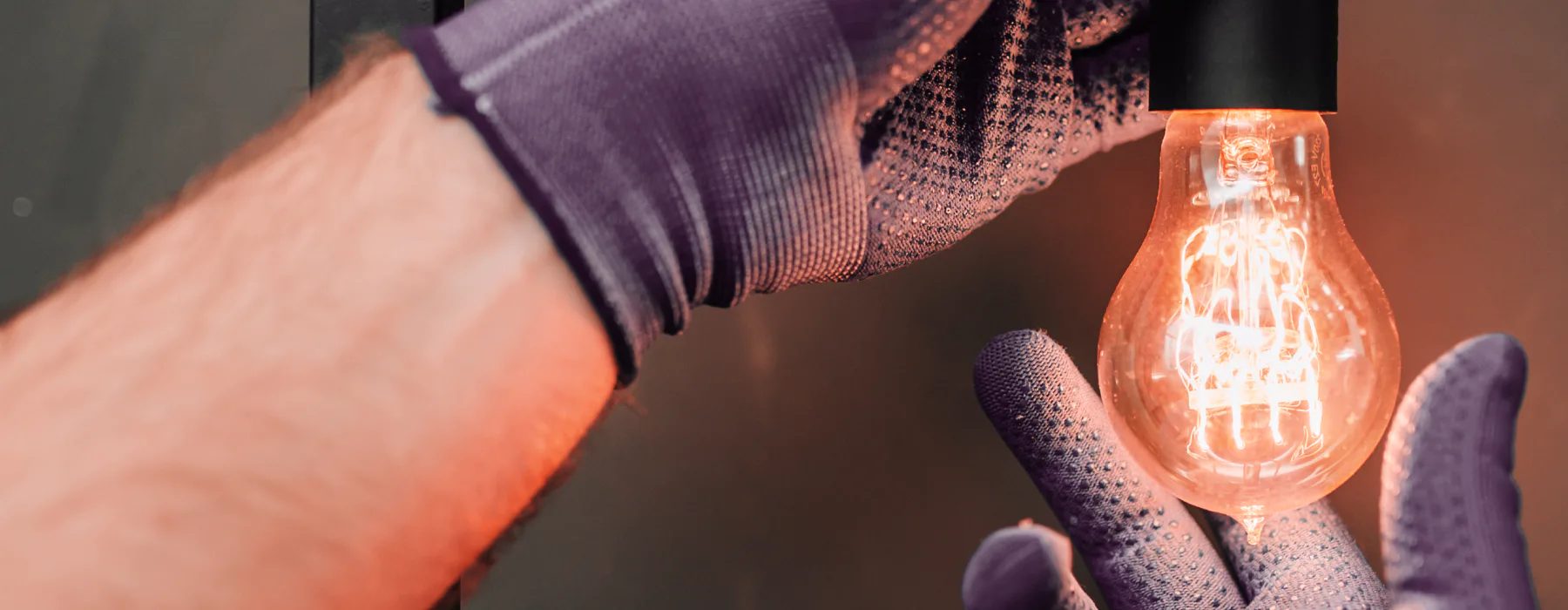 A panoramic, close-up image showing a person wearing purple, textured work gloves screwing in a lit, vintage-style Edison lightbulb with a glowing orange filament into a black socket.