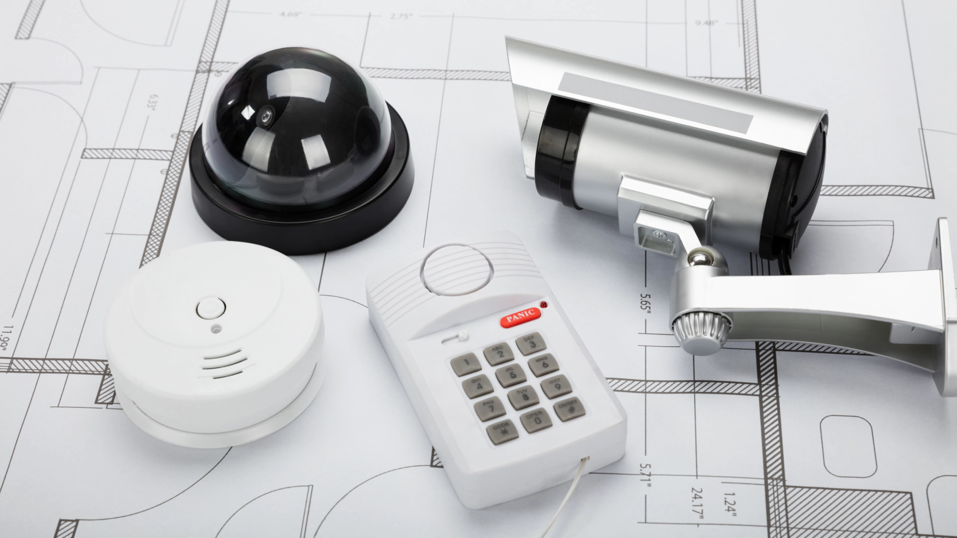 A collection of home security system components laid out on a set of architectural blueprints, including a black dome camera, a silver bullet camera, a white smoke detector, and a security keypad with a red panic button.